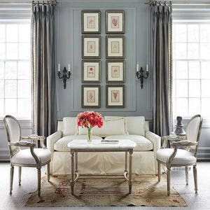 A vertical display of eight botanical prints arranged side-by-side in two vertical columns on a gray wall between two tall windows with drapes. Dark wood frames with cream colored mats. Black double sconces on each side of the display.