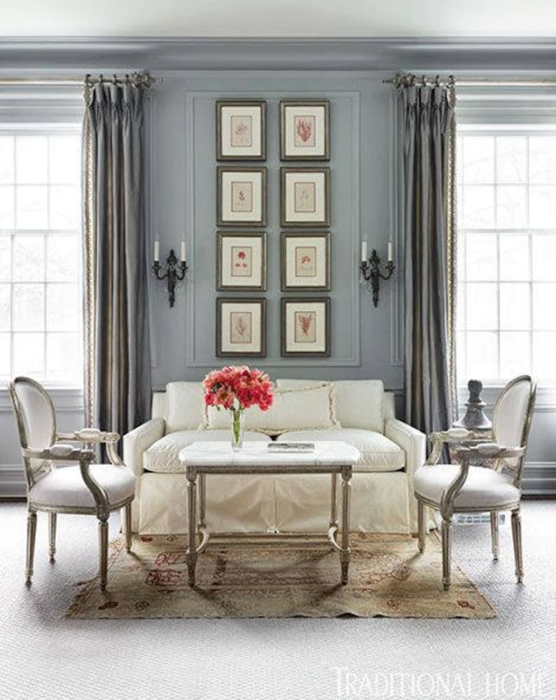 A vertical display of eight botanical prints arranged side-by-side in two vertical columns on a gray wall between two tall windows with drapes. Dark wood frames with cream colored mats. Black double sconces on each side of the display.