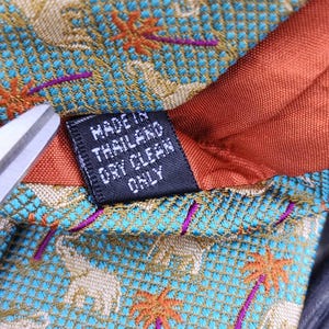 May include: Close-up of a patterned tie with a teal background featuring elephants and palm trees in orange and purple. The tie has a rust-colored lining and a tag that reads "MADE IN THAILAND DRY CLEAN ONLY."