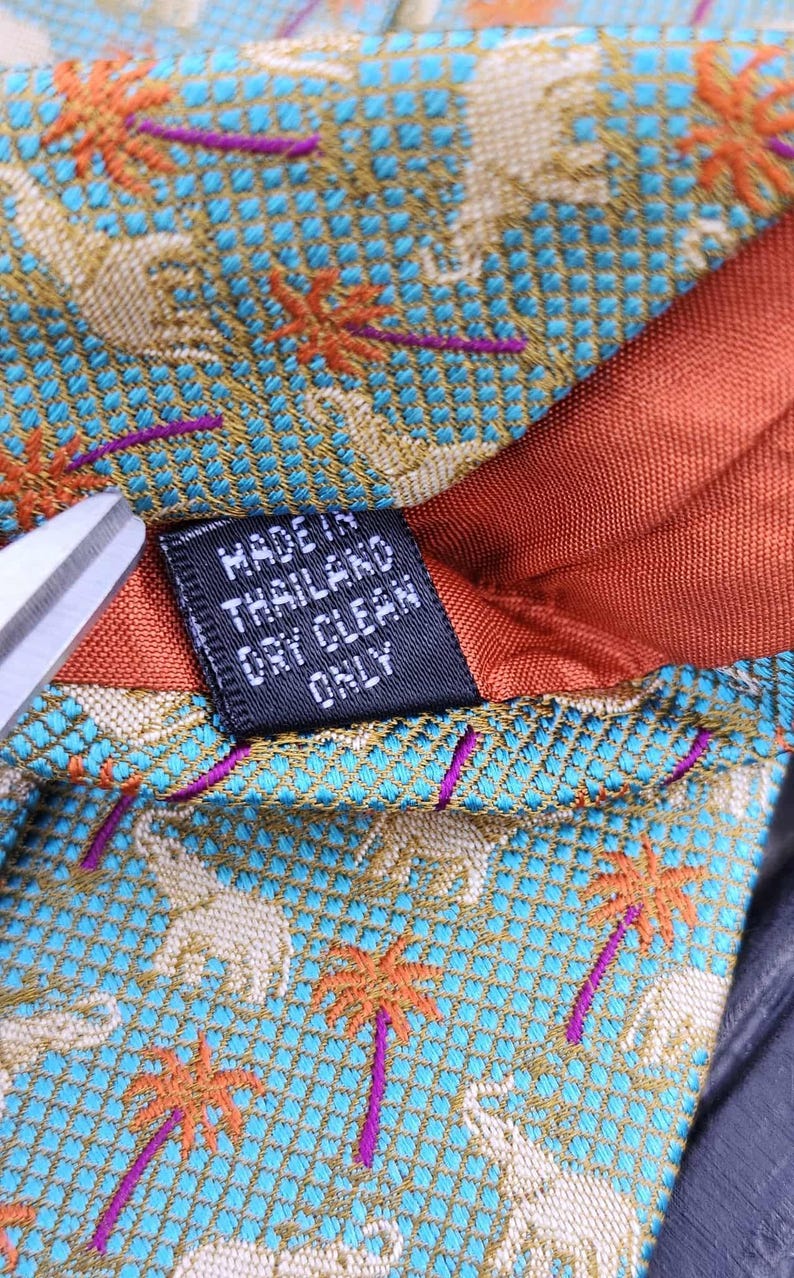 May include: Close-up of a patterned tie with a teal background featuring elephants and palm trees in orange and purple. The tie has a rust-colored lining and a tag that reads "MADE IN THAILAND DRY CLEAN ONLY."