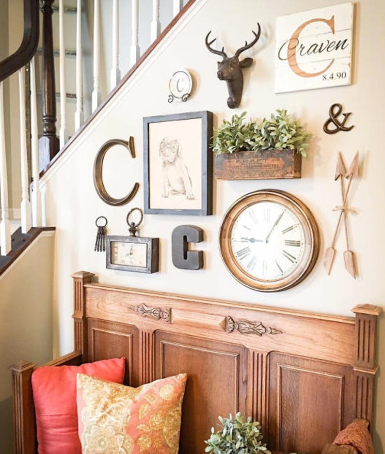 Small wallspace below a set of stairs is decorated with a carved wooden bench and throw pillows. Above the bench is an asymmetric gallery display with framed and unframed artwork, a clock, wooden monograms, and other knickknacks hanging on the wall.