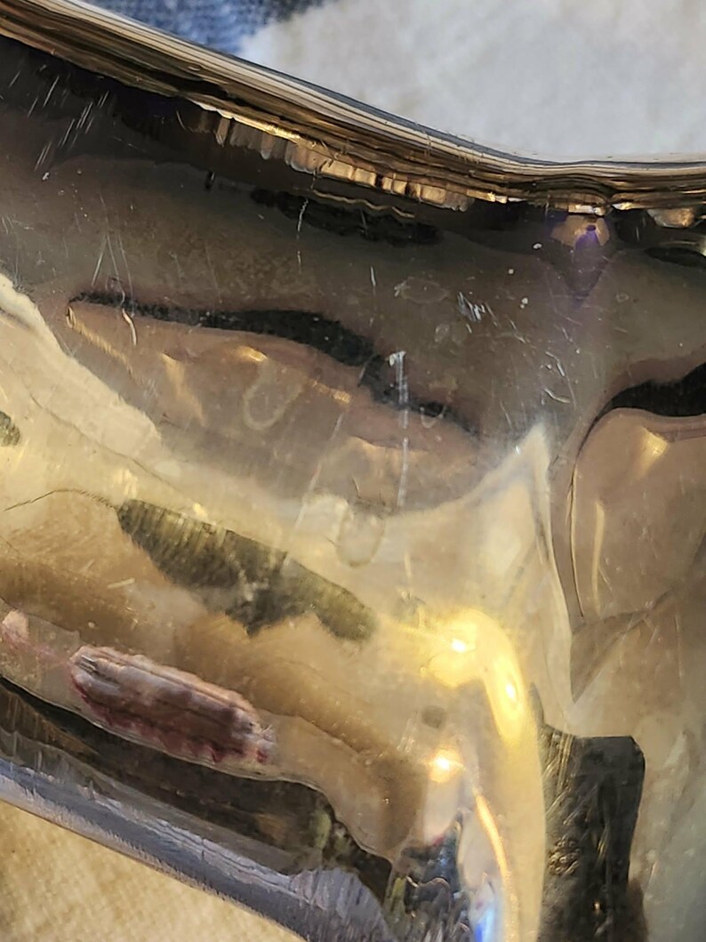 May include: Close-up of a shiny, silver-colored object with a curved shape. The surface reflects light, showing distorted reflections and minor scratches. The object appears to be made of metal, possibly a decorative piece.