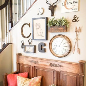 Small wallspace below a set of stairs is decorated with a unique asymmetric gallery-style display with framed and unframed artwork, a clock, wooden monograms, and other knickknacks. A carved wooden bench with throw pillows sits below the arrangement.