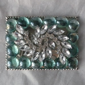 Puede incluir: Una hebilla de cintur&oacute;n plateada con un dise&ntilde;o de diamantes de imitaci&oacute;n transparentes y cuentas de vidrio de color verde azulado.