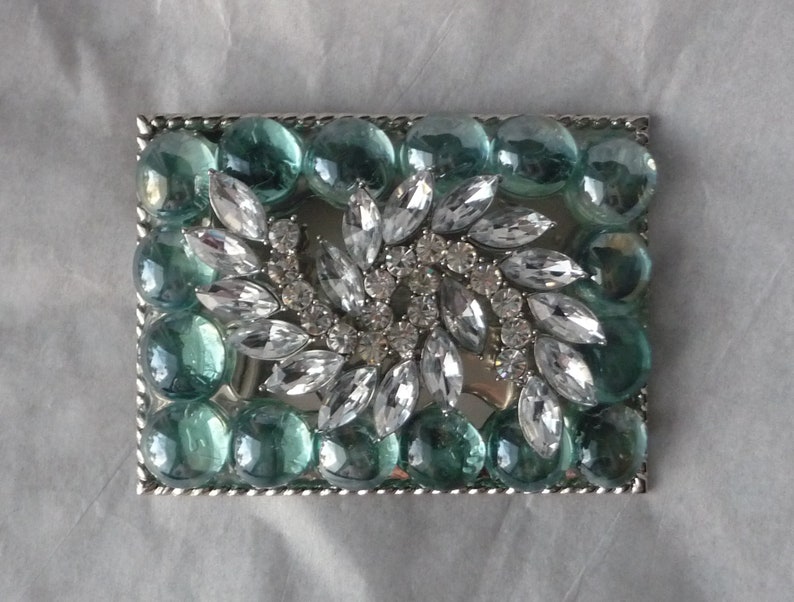 Puede incluir: Una hebilla de cintur&oacute;n plateada con un dise&ntilde;o de diamantes de imitaci&oacute;n transparentes y cuentas de vidrio de color verde azulado.