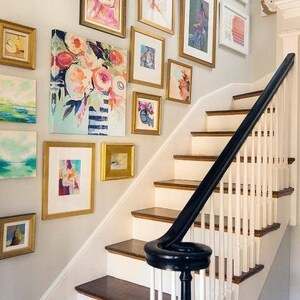 May include: A white staircase with a black handrail and a wall decorated with a gallery of framed artwork in gold frames. The artwork features abstract paintings, landscapes, and floral designs.