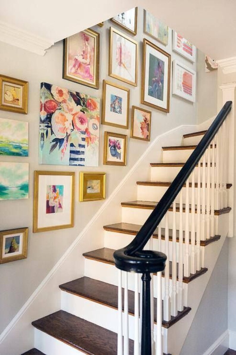 May include: A white staircase with a black handrail and a wall decorated with a gallery of framed artwork in gold frames. The artwork features abstract paintings, landscapes, and floral designs.