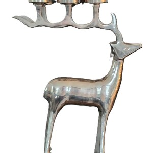 May include: Silver-colored metal deer sculpture with three candle holders on its antlers. The deer has a sleek, modern design, with a polished finish. The sculpture is a decorative item.