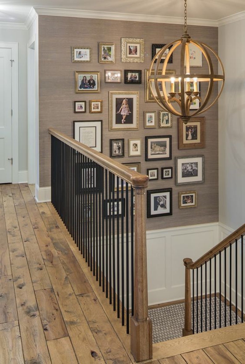 May include: A wooden staircase with a black metal railing leads up to a wall decorated with framed photos and a large, round, gold-toned chandelier.