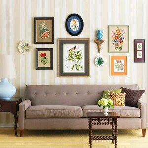 A living room with a gallery-style display of framed floral prints of various shapes and sizes, decorative plates and other wall hangings placed in appealing asymmetrical arrangement on white and beige striped wallpaper above a light brown couch.