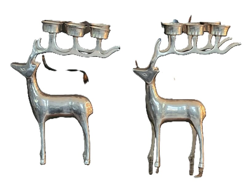 May include: Two silver-colored reindeer candle holders. Each reindeer has a rack of antlers that hold four candle cups. The reindeer are standing upright with their heads held high. The candle holders are a festive decoration.