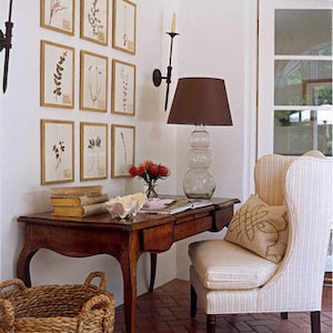Classic style of nine gold-framed botanical leaf prints in a symmetrical gallery-style arrangement of three rows on a white wall above a writing desk with an upholstered wing back chair. Black wrought iron sconces flank the framed prints.