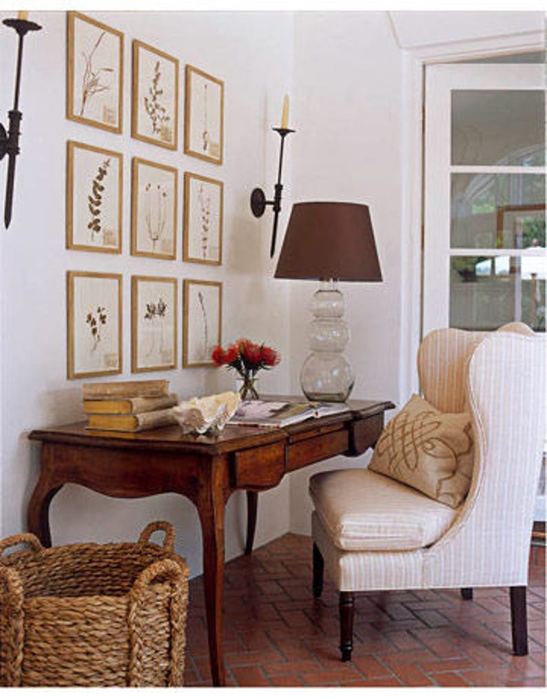 Classic style of nine gold-framed botanical leaf prints in a symmetrical gallery-style arrangement of three rows on a white wall above a writing desk with an upholstered wing back chair. Black wrought iron sconces flank the framed prints.