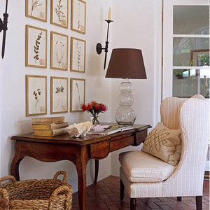 A symmetrical, gallery-style wall display with nine gold-framed botanical leaf prints arranged in 3 rows on a white wall above a writing desk with an upholstered wing back chair. Black wrought iron sconces flank the framed prints.