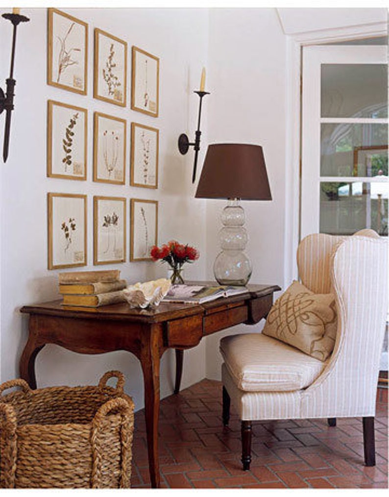 A symmetrical, gallery-style wall display with nine gold-framed botanical leaf prints arranged in 3 rows on a white wall above a writing desk with an upholstered wing back chair. Black wrought iron sconces flank the framed prints.