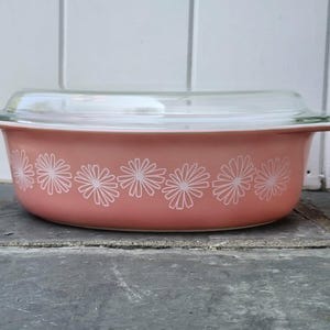 May include: An oval-shaped pink casserole dish with a clear glass lid. The exterior of the dish features a repeating white floral pattern consisting of stylized daisy-like flowers arranged in a horizontal row around the center of the vessel.