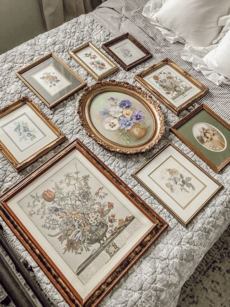 May include: A collection of framed floral prints and paintings in various sizes and styles, arranged on a white and gray quilted bedspread. The frames are all gold or brown and have a vintage aesthetic.