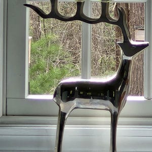 May include: A silver-colored deer-shaped candle holder with three candle cups. The deer has a sleek, modern design and stands in front of a window with a view of trees. The candle holder is made of metal and has a reflective surface.