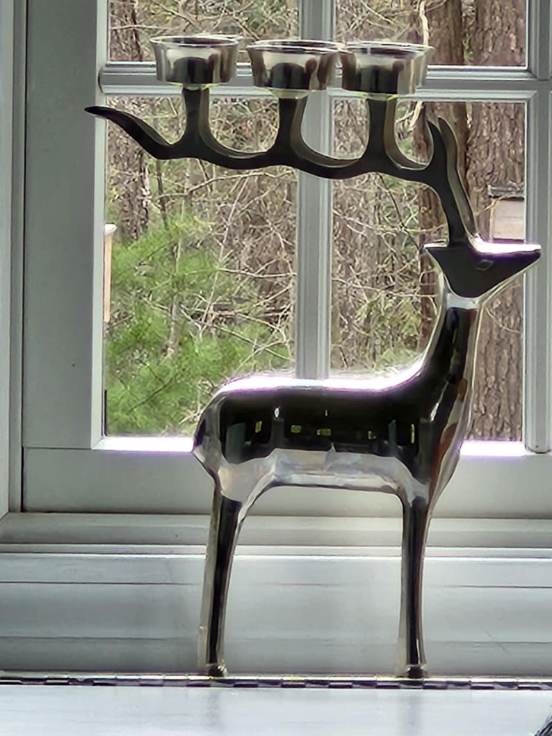 May include: A silver-colored deer-shaped candle holder with three candle cups. The deer has a sleek, modern design and stands in front of a window with a view of trees. The candle holder is made of metal and has a reflective surface.