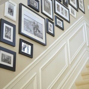May include: A white wall with black picture frames of various sizes, displaying black and white photos. The frames are arranged in a staggered pattern, creating a visually appealing gallery wall. The wall is adjacent to a white staircase with a beige carpet.