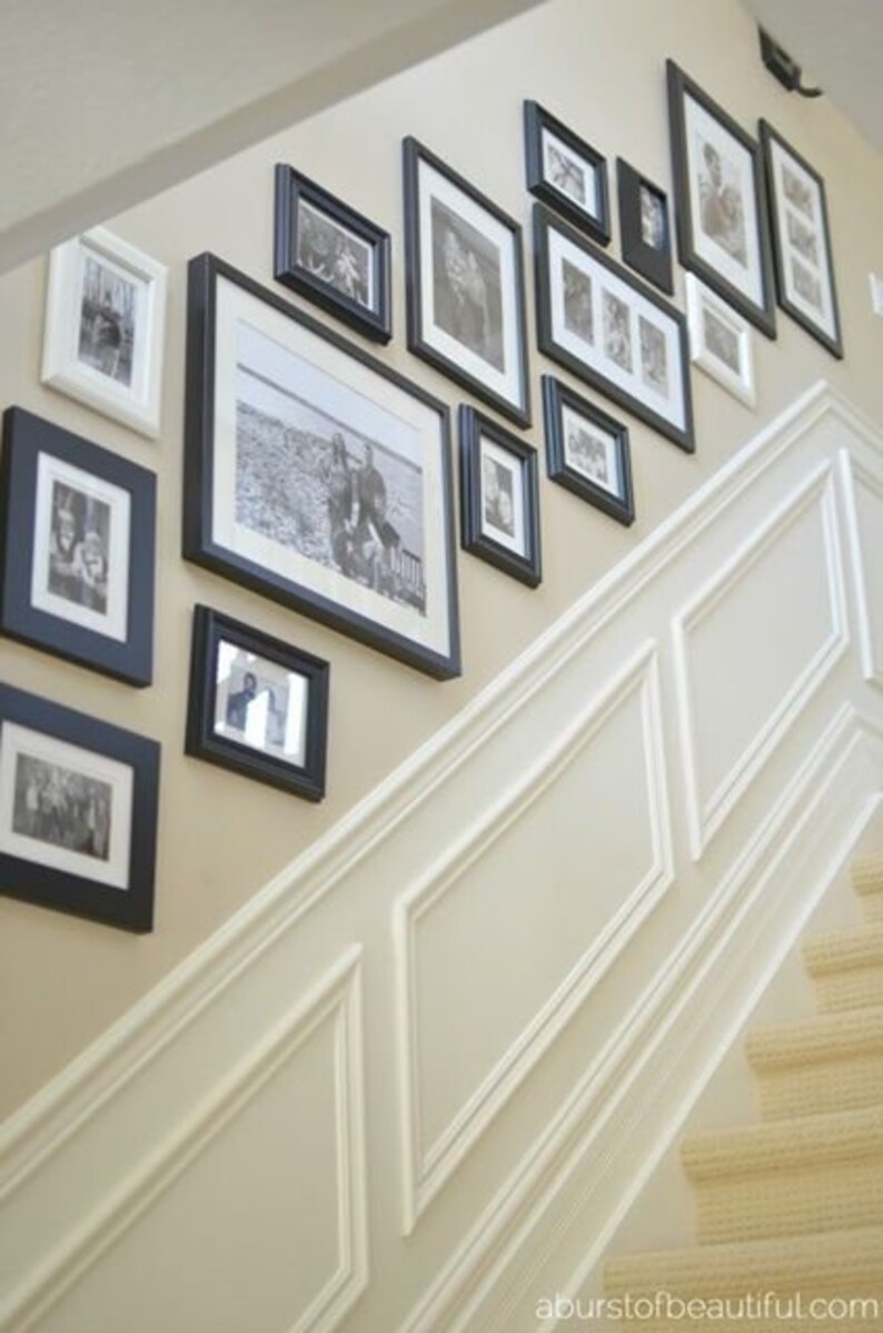 May include: A white wall with black picture frames of various sizes, displaying black and white photos. The frames are arranged in a staggered pattern, creating a visually appealing gallery wall. The wall is adjacent to a white staircase with a beige carpet.