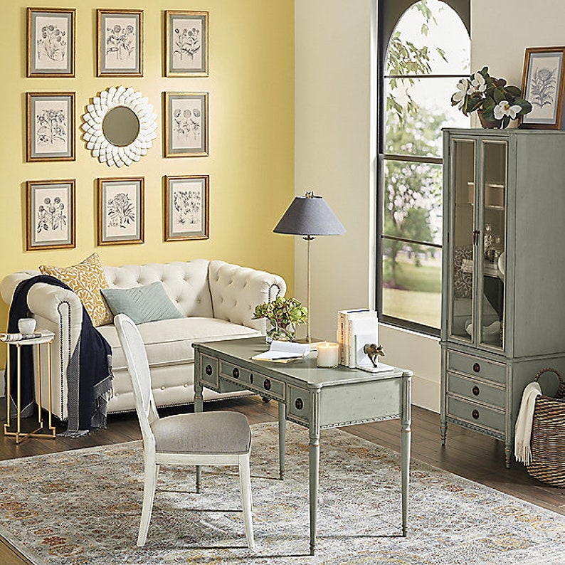 May include: A light green desk with four drawers and turned legs sits in a home office with a white sofa, a white chair, and a tall cabinet with glass doors. The room has a yellow wall with framed botanical prints and a white mirror.