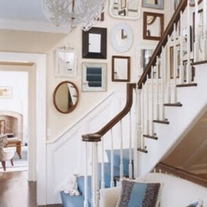 Example of a foyer with a gallery-style display of numerous mirrors of various shapes and sizes in white, gold or black frames hung on a light beige wall next to a set of stairs.