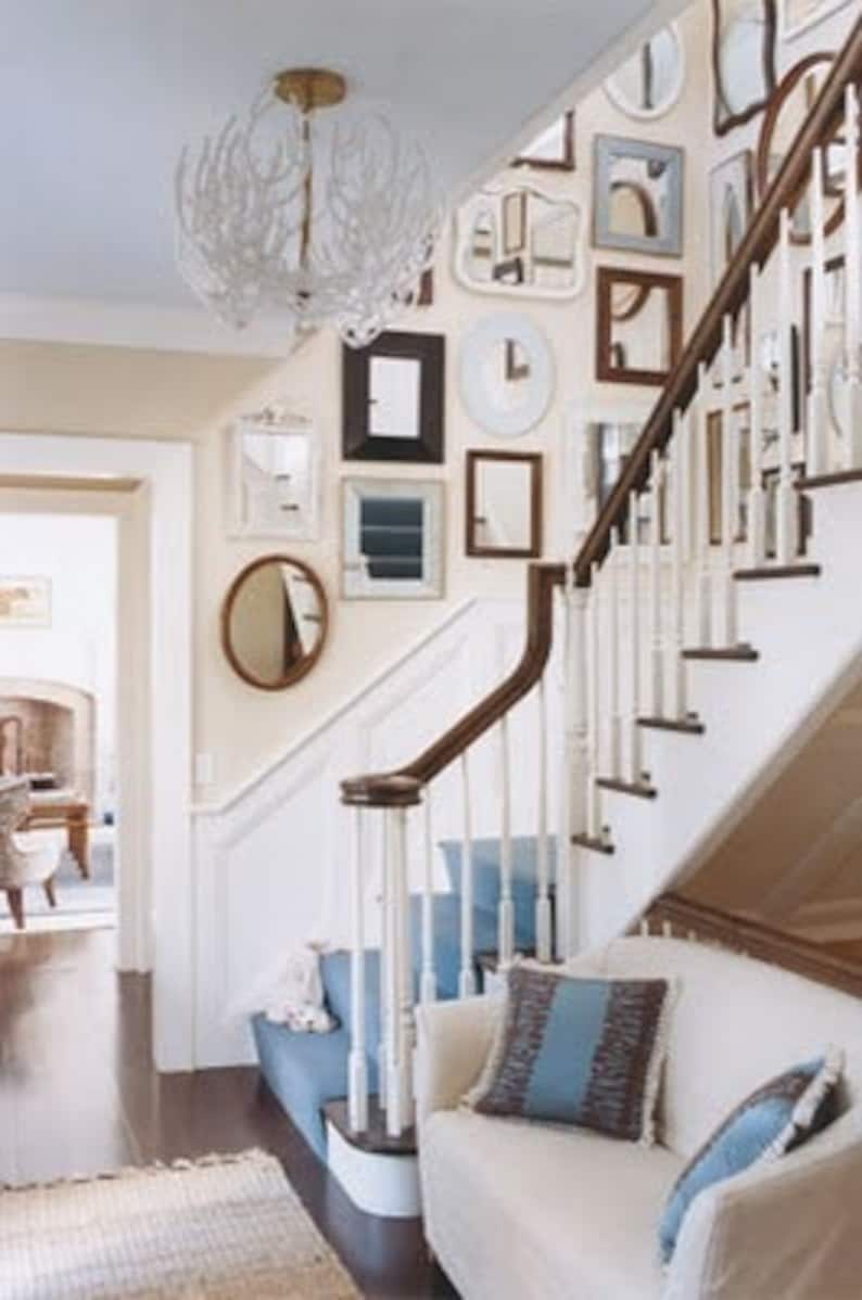 Example of a foyer with a gallery-style display of numerous mirrors of various shapes and sizes in white, gold or black frames hung on a light beige wall next to a set of stairs.