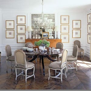 Unique example of a dining room with a symmetrical, gallery-style display on three adjacent white walls. The small, square art pieces are set off with large off-white mats and thin gold frames. Dark table and dark herringbone wood floor.