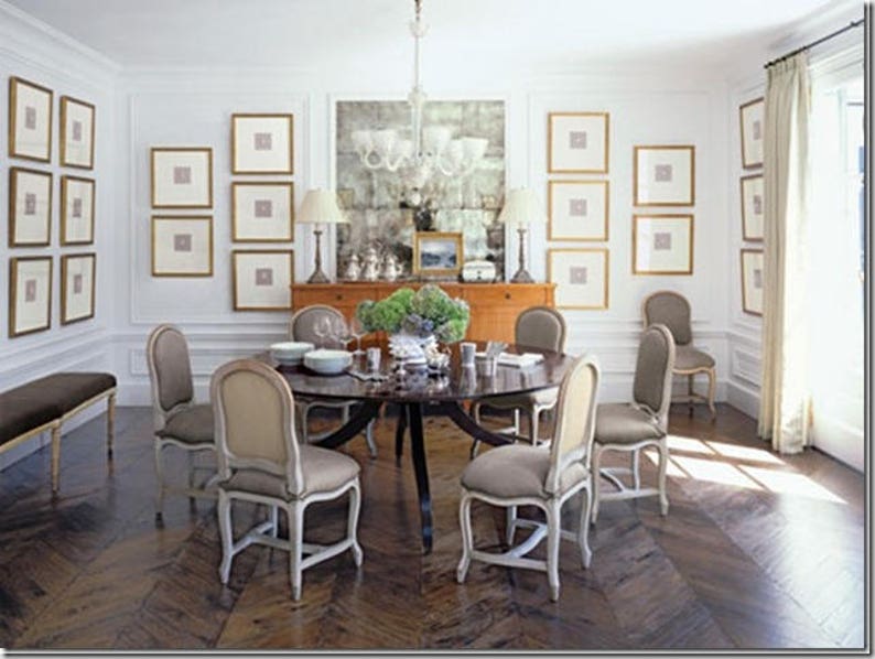 Unique example of a dining room with a symmetrical, gallery-style display on three adjacent white walls. The small, square art pieces are set off with large off-white mats and thin gold frames. Dark table and dark herringbone wood floor.
