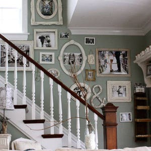 A gallery-style display of white-framed artwork on a green wall next to a set of stairs. The art includes framed prints and photos in various shapes and sizes, mirrors, and other miscellaneous pieces placed in an appealing arrangement.