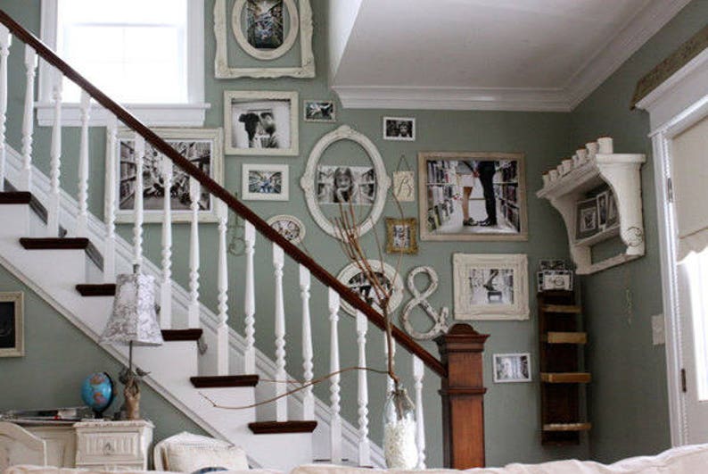 A gallery-style display of white-framed artwork on a green wall next to a set of stairs. The art includes framed prints and photos in various shapes and sizes, mirrors, and other miscellaneous pieces placed in an appealing arrangement.