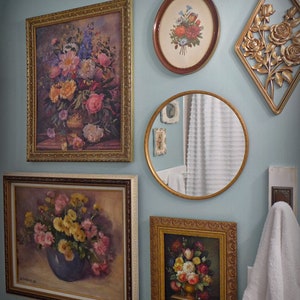 May include: A bathroom wall decorated with framed floral paintings and a round mirror. The paintings feature various floral arrangements in vases, with a mix of pink, yellow, and red flowers. The mirror reflects a white bathroom with a white towel hanging on a hook.