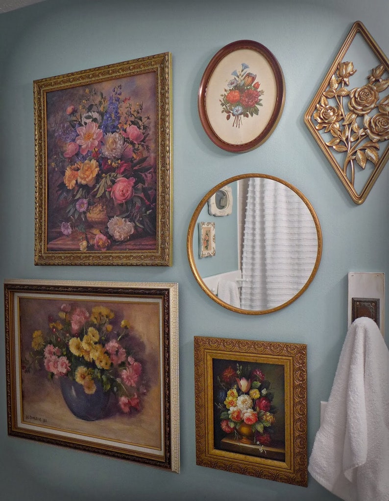 May include: A bathroom wall decorated with framed floral paintings and a round mirror. The paintings feature various floral arrangements in vases, with a mix of pink, yellow, and red flowers. The mirror reflects a white bathroom with a white towel hanging on a hook.