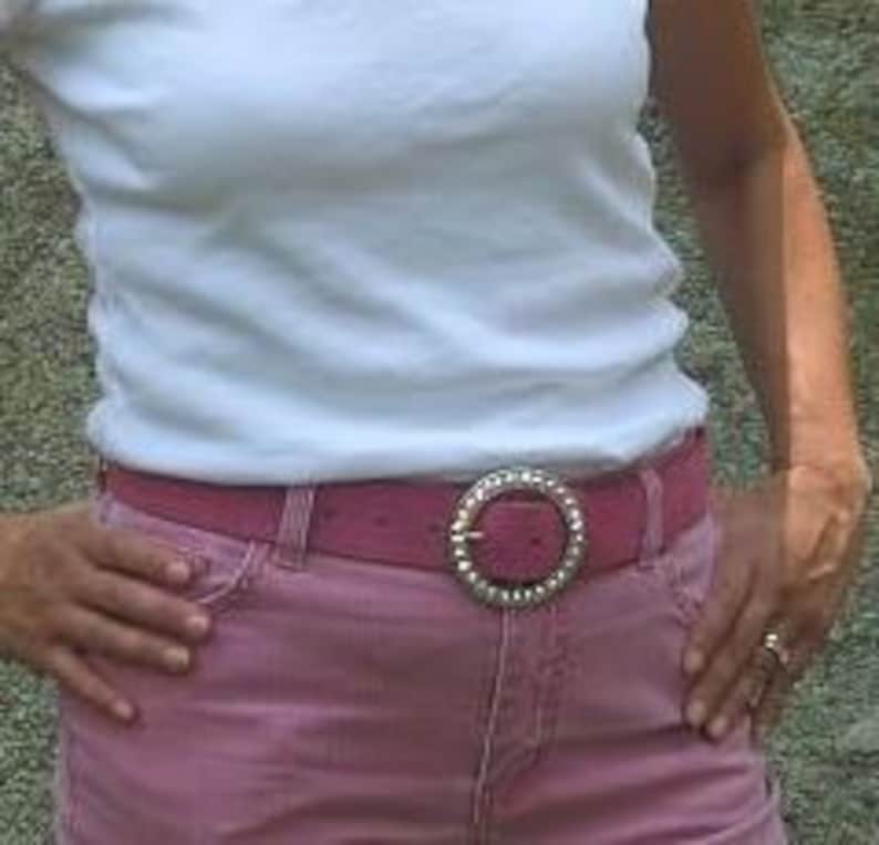 Puede incluir: Un cintur&oacute;n rosa con una hebilla plateada grande con un c&iacute;rculo de diamantes de imitaci&oacute;n. El cintur&oacute;n se lleva sobre unos vaqueros rosas.
