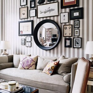 Striking example of an asymmetrical gallery-style display that includes black-framed art and photos of various shapes and sizes, a convex mirror, and other wall hangings placed in an appealing arrangement on tan striped wallpaper above a couch.
