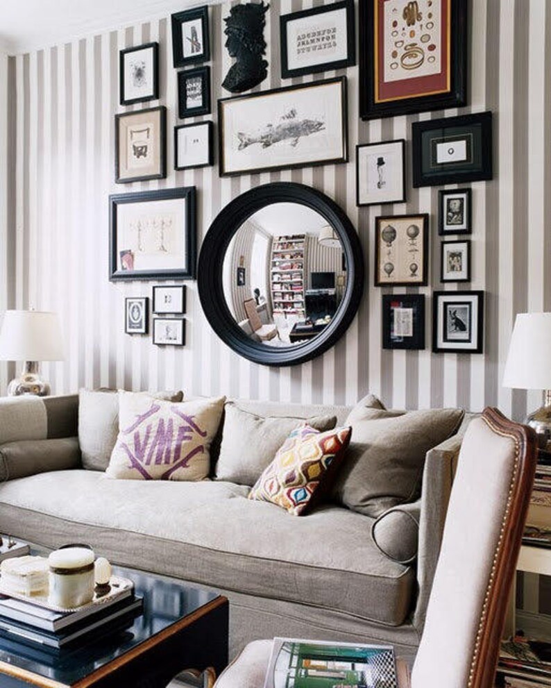 Striking example of an asymmetrical gallery-style display that includes black-framed art and photos of various shapes and sizes, a convex mirror, and other wall hangings placed in an appealing arrangement on tan striped wallpaper above a couch.