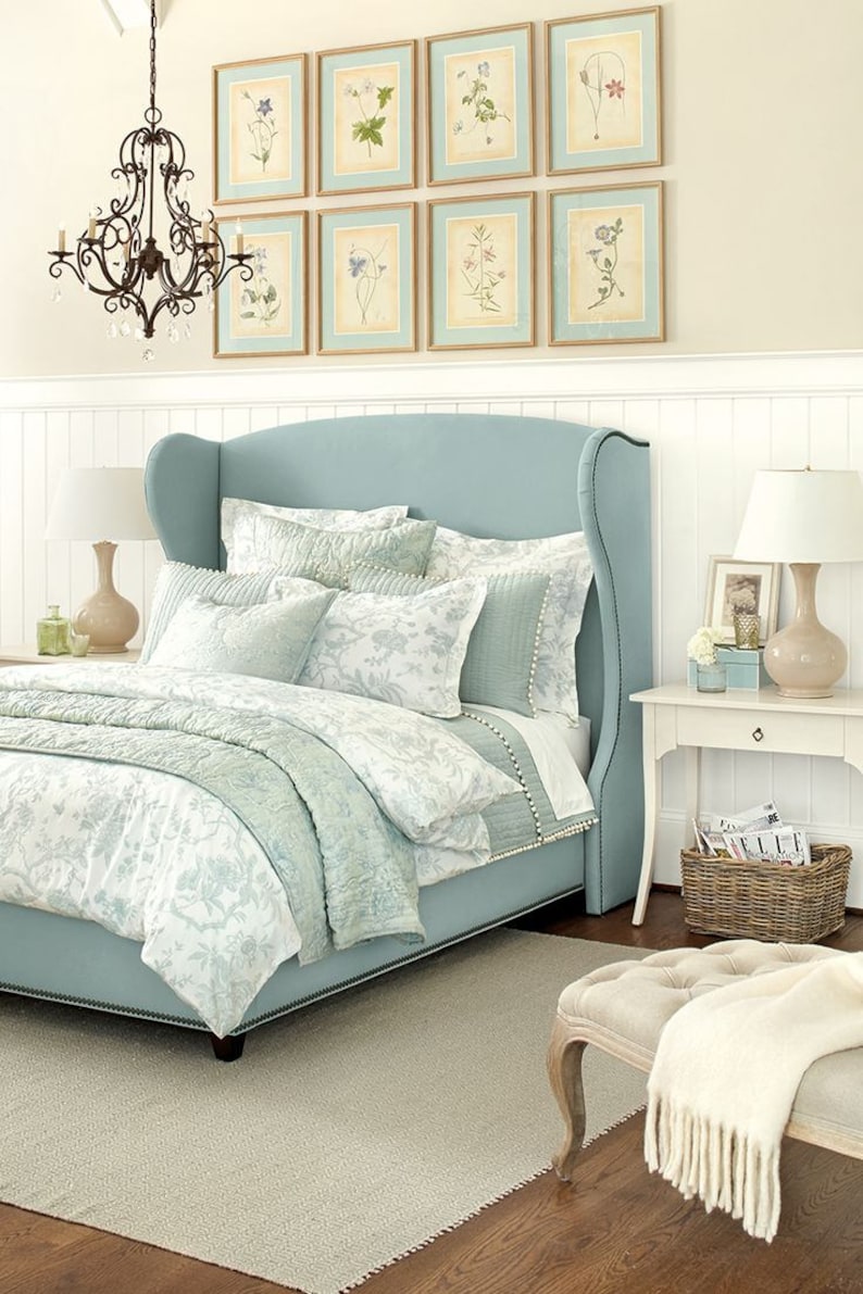 Puede incluir: Una cama tapizada de color azul claro con una cabecera alta y un edred&oacute;n con estampado floral blanco y azul claro. La cama est&aacute; en un dormitorio con z&oacute;calos blancos y una alfombra blanca. Hay estampados bot&aacute;nicos enmarcados en la pared sobre la cama.