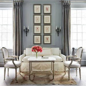 Handsome example of a vertical display of eight botanical prints arranged in two vertical columns on a gray wall between two tall windows with drapes. Dark wood frames with cream colored mats. Black double sconces on each side of the display.