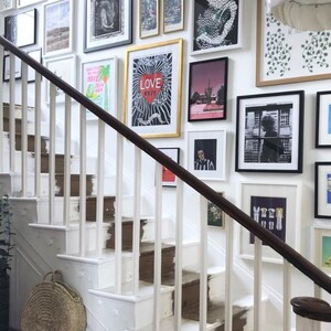 May include: A white staircase with a wooden handrail. The wall is decorated with a gallery wall of framed artwork, including a black and white photo of a person, a poster with the word "LOVE" in red, and a poster with a snake design.