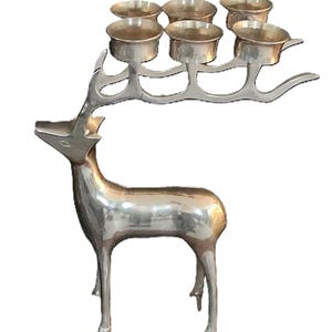 May include: A silver-colored metal deer sculpture with six candle holders on its antlers. The deer is standing, with a polished finish, and the candle holders are small, round cups.