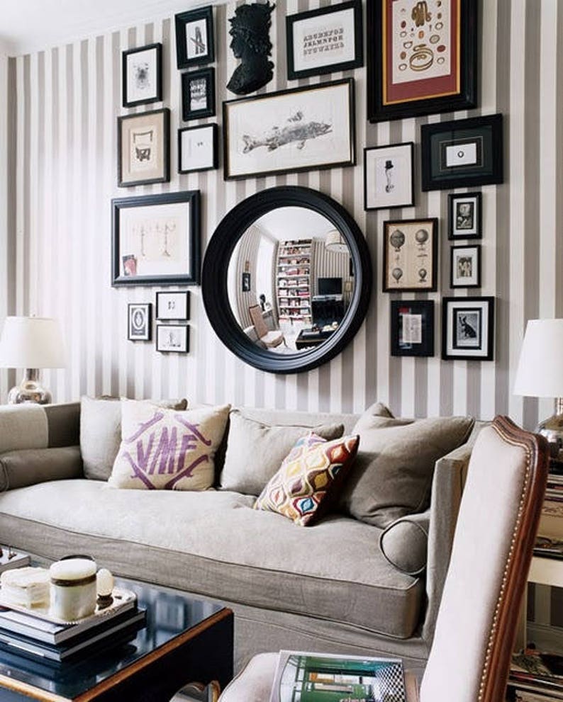 Example of a living room gallery-style wall display that includes black-framed art and photos of various shapes and sizes, a convex mirror, and other wall hangings placed in an appealing arrangement on tan striped wallpaper above a couch.