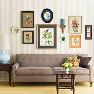 An asymmtrical gallery-style wall display with framed floral prints of various shapes and sizes, decorative plates and other wall hangings placed in an appealing arrangement on white and beige striped wallpaper over a light brown couch.