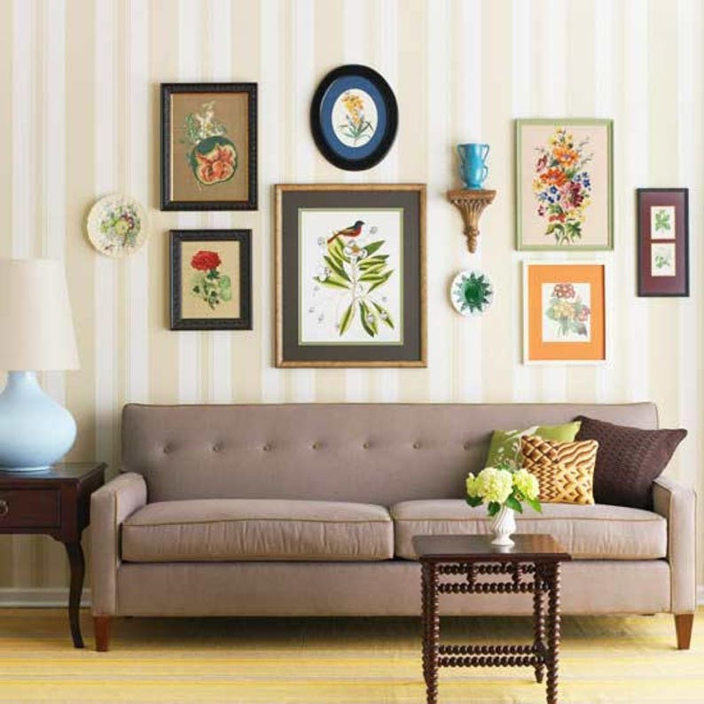 An asymmtrical gallery-style wall display with framed floral prints of various shapes and sizes, decorative plates and other wall hangings placed in an appealing arrangement on white and beige striped wallpaper over a light brown couch.