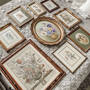 May include: A collection of framed floral prints and paintings in various sizes and styles, arranged on a white and gray quilted bedspread. The frames are all gold or wood and have a vintage aesthetic.