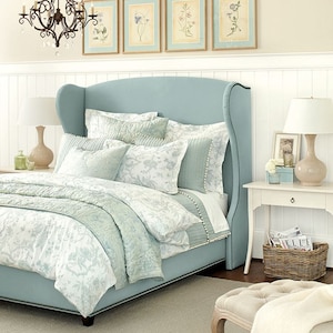 May include: A light blue upholstered bed with a high headboard and a white and light blue floral patterned duvet cover. The bed is in a bedroom with white wainscoting and a white rug. There are framed botanical prints on the wall above the bed.