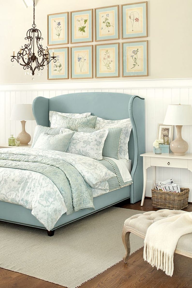 May include: A light blue upholstered bed with a high headboard and a white and light blue floral patterned duvet cover. The bed is in a bedroom with white wainscoting and a white rug. There are framed botanical prints on the wall above the bed.