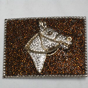 Puede incluir: Un broche de cabeza de caballo de plata y oro con un fondo de cuentas marrones. La cabeza de caballo est&aacute; decorada con diamantes de imitaci&oacute;n.