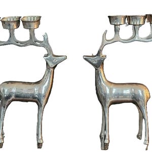 May include: Two silver-colored deer candle holders. Each deer has a rack of antlers that hold three candle cups. The deer are standing upright with their heads facing each other. The candle holders are made of metal.