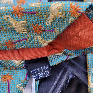 May include: Close-up of a silk tie with a teal background featuring a pattern of elephants and palm trees in gold and orange. The tie has a red lining and a label that reads "100% Silk".